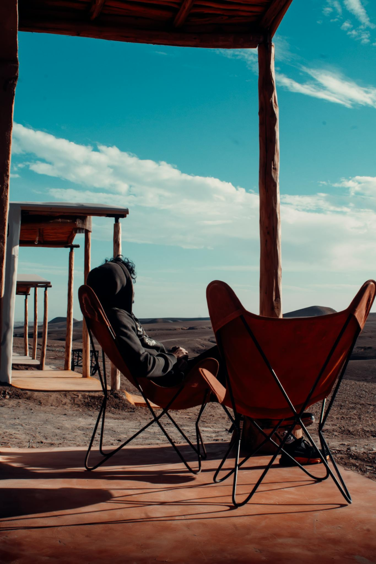 A man sits on a porch overlooking the arid desert landscape of Agafay, Morocco, under a sunlit sky.