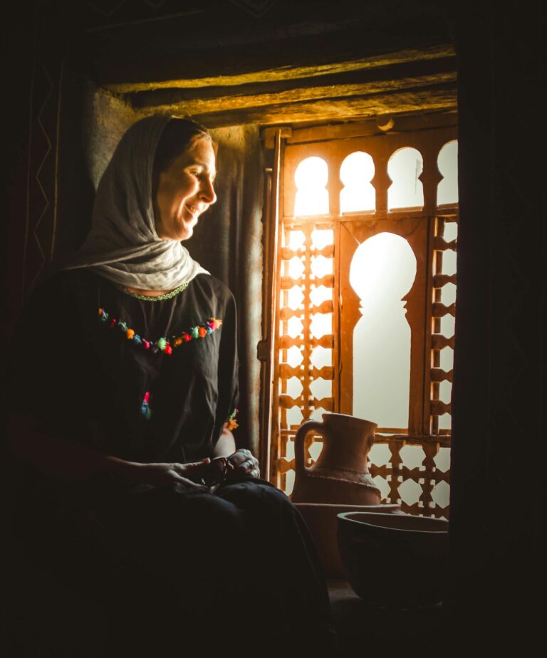 A woman in traditional Moroccan attire sits by an ornately patterned window in Agdz, Morocco.